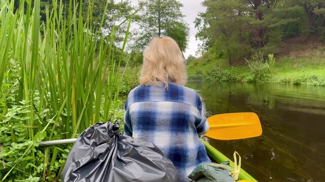 Woman sitting in canoe beside tall riverbank reeds with yellow paddle resting at her side as calm Irbe River in Latvia flows forward in soft overcast light
