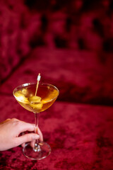 A close-up shot of a hand holding a classic cocktail, possibly a dry martini or gimlet, in a coupe glass with green olives as garnish, against a deep red velvet background