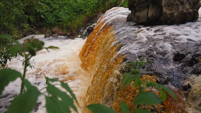 Brown water with white foam rushes over a rocky waterfall in a lush green forest, creating a powerful and dynamic natural scene. The current flows rapidly downstream in cascades. Belarus Vyata River