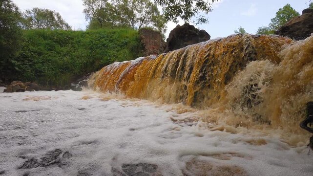 A powerful stream of murky brown water cascades over the rocks in the lush green forest, creating white foam as it flows down the river and highlighting the raw force of nature in the wild. Belarus Vy