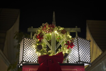 Glowing oversized Christmas wreath adorned with red poinsettias, berries, and twinkling fairy lights, framed by wooden lattice panels and festive red banner. Nighttime holiday magic against dark sky e