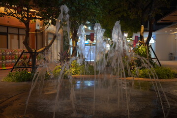 Enchanting nighttime water fountain display with illuminated arcs spraying gracefully among lush tropical trees and plants. Reflective wet tiles shimmer beside modern shopping mall facade with red lan