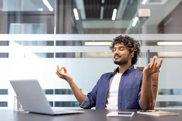 Fotobehang Lotusbloem Happy young Indian man meditating relaxed in the office at his desk, sitting in lotus position with eyes closed  © Tetiana
