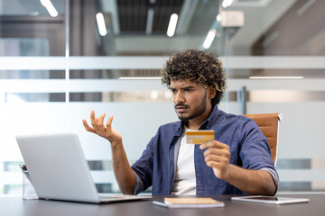 Frustrated young Indian man found an error and problem with his bill and credit card which he holds in his hand and looks at his laptop