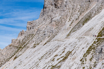 Hiking trail with some hikers at base of Monte Paterno mountain near north side of Tre Cime di Lavaredo peaks also called Drei Zinnen with white clouds in Dolomite Alps, near Cortina d'Ampezzo, Italy