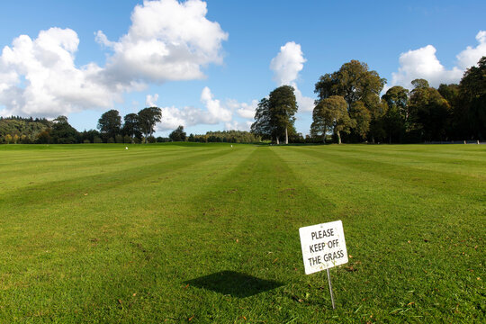 Low angle view over two toned striped grass of a golf course in Ireland with in front a sign to keep off the grass with trees in background against a white clouded blue sky