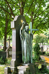 Große Jesusfigur mit einem Kreuz in der Hand steht vor einem großen Grabstein mit einer strahlenden Sonne auf dem katholischen Friedhof in Kempten © Blende8