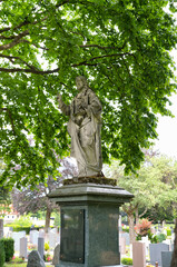 sehr alte stark verwitterte Jesusstatue auf einem Sockel unter einem Baum auf dem Waldfriedhof in Memmingen © Blende8
