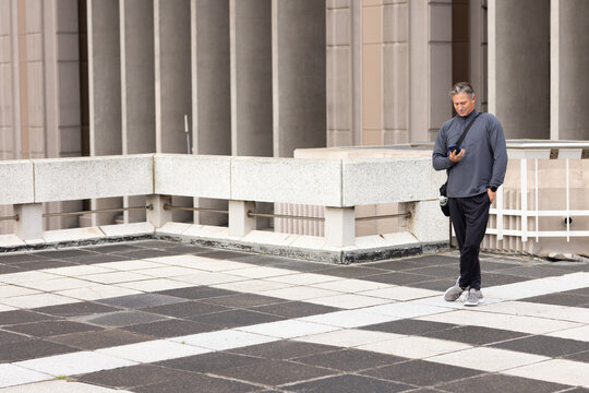 Middle-aged man standing on rooftop terrace holding smartphone and crossbody camera bag, copy space