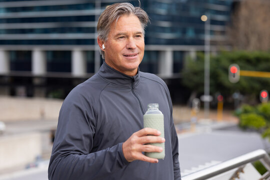 Man wearing gray jacket and earbuds standing on pedestrian walkway holding water bottle by railing