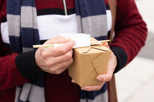 Mature adult man standing on sidewalk placing chopsticks on brown food container wearing scarf