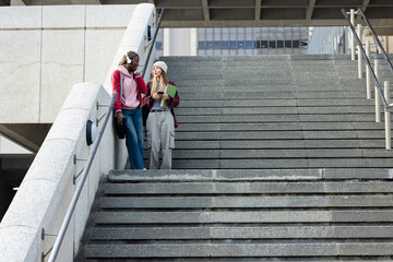 Diverse female friends chatting on urban steps with smartphone headphones coffee cup notebook