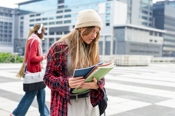 Diverse female students near crosswalk in plaza holding notebooks and listening with headphones