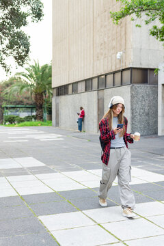 Woman walking plaza beside concrete wall, scrolling smartphone and holding coffee cup, copy space