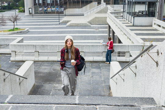 Female student ascending stairs with backpack holding coffee cup and smartphone at campus courtyard