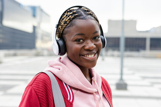 African American woman standing on rooftop wearing pink hoodie, red varsity jacket, headphones