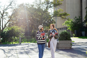 Diverse female students walking on campus walkway chatting while holding notebooks and smartphone