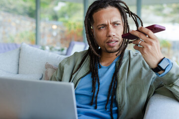 Middle-aged African American man talking on smartphone while typing on laptop on sofa, copy space