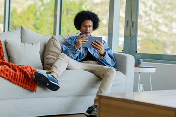African American man sitting on gray sofa holding tablet focusing on screen in modern living room
