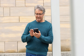 Male wearing teal sweater and eyeglasses, leaning against column holding smartphone outdoors