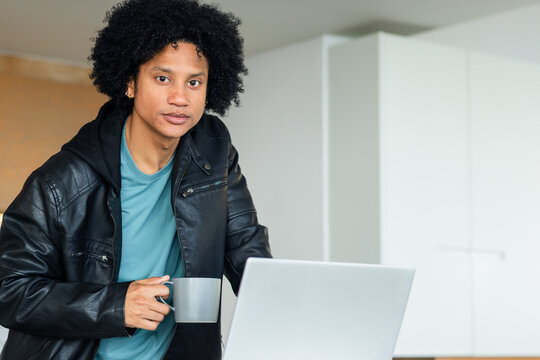 Mid adult African American man leaning on modern kitchen counter holding mug by laptop