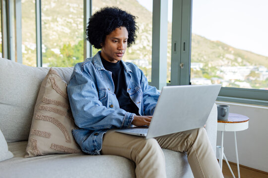 African American man typing on laptop on sofa with coffee mug in living room overlooking hillside