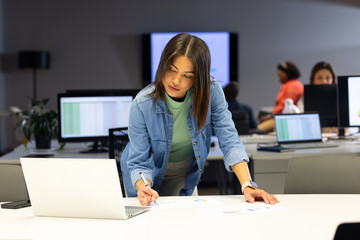 Indian woman leaning forward at desk in open-plan office reviewing charts with pen and open laptop