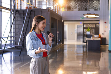 Asian speaker standing with microphone and gesturing in lobby with staircase, badge, grid windows