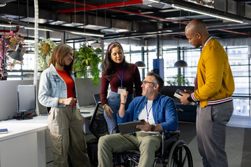 Diverse coworkers gathering around man in wheelchair collaborating on tablet in open plan office