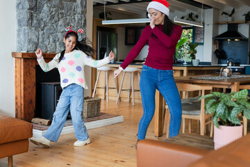 Mother and Indian daughter dancing on wood floor in home wearing Santa hat and reindeer headband