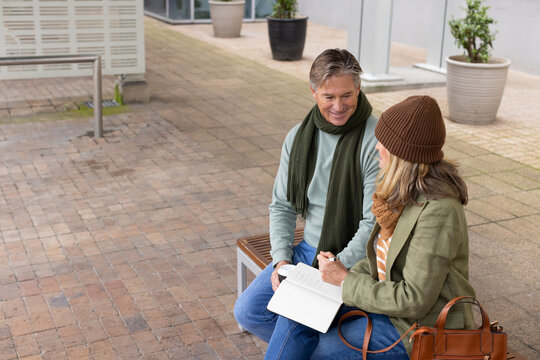 Colleagues sitting on wooden bench in courtyard writing notes with notebook and pen, copy space