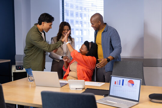 Diverse coworkers high-fiving at office analyzing laptop charts with desk phone and water glass