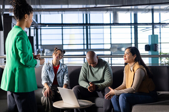 Diverse coworkers discussing ideas on sofa around coffee table with laptop and mug in office lounge - Powered by Adobe