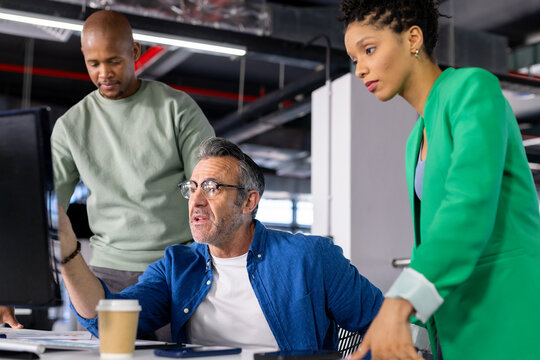 Diverse coworkers pointing at monitor and reviewing work at office desk with coffee cup, papers