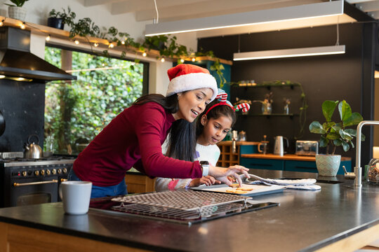 Diverse mother and daughter wearing Santa hats cutting cookies on baking sheet on kitchen island