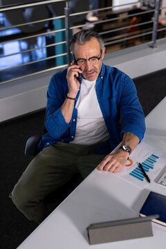 Businessman calling on smartphone while pointing at bar chart at desk in modern office