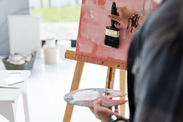 Indian man applying red strokes on canvas on easel using flat paintbrush and palette in studio