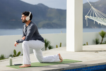 Indian man lunging on yoga mat at poolside terrace wearing athletic top and pants, copy space
