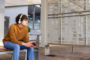Man holding red smartphone on bench in shelter wearing sweater with headphones by stacked notebooks