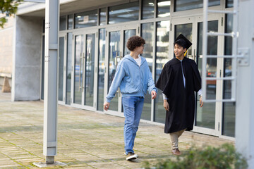 Diverse male students crossing walkway by support post outside glass doors wearing mortarboard gown