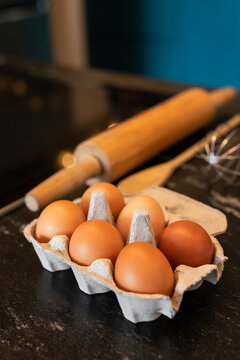 Six brown eggs resting in grey cardboard tray on countertop, with rolling pin and whisk blurred