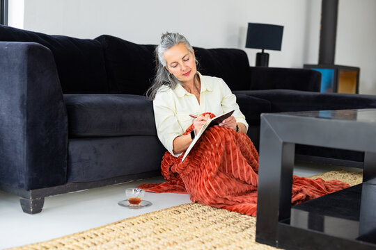 Senior woman writing in notebook on living room floor wrapped in rust-red blanket with tea cup