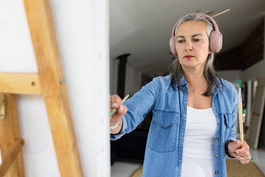 Female painter painting canvas on easel in studio using two brushes wearing headphones, copy space