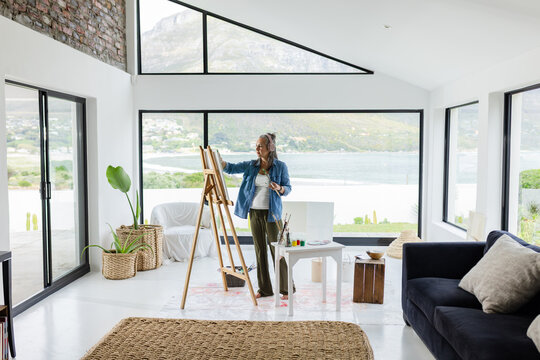 Female painter painting canvas at easel palette in living room overlooking water and mountains