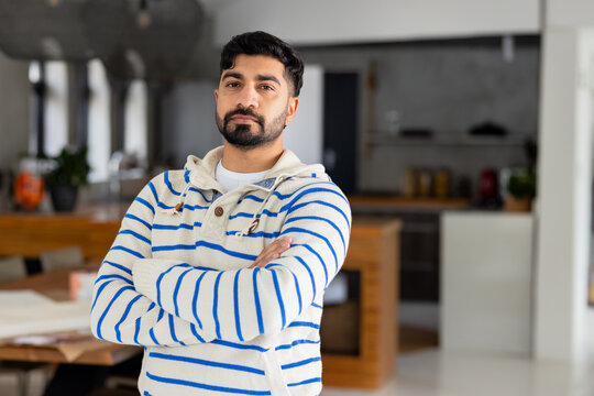Indian man standing with arms crossed in modern kitchen beside table with papers, mug and plant
