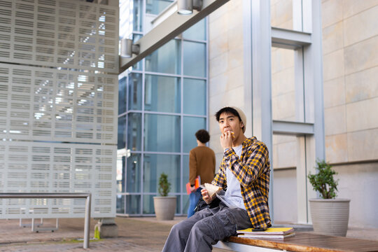 Asian male student eating pastry on wooden bench at campus courtyard beside notebooks and paper bag