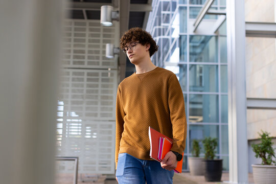 Male student walking along building corridor holding colorful notebooks and checking watch
