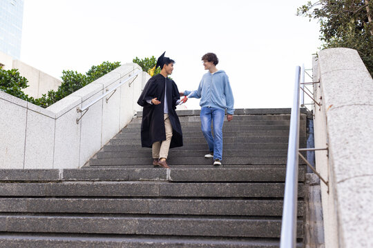 Diverse male friends walking down campus steps in mortarboard cap handing rolled ribboned diploma