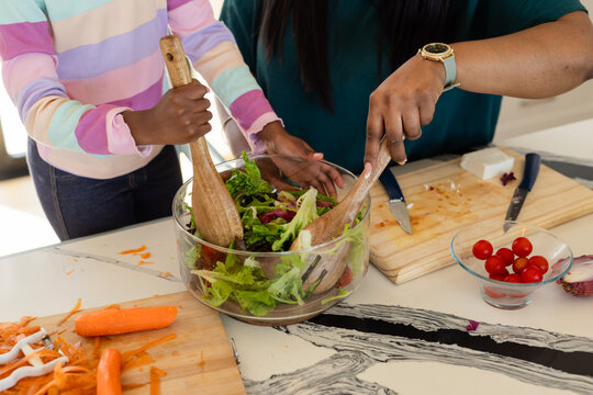 African American mother and daughter mixing salad in glass bowl at counter with wooden spoons
