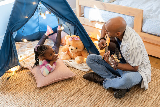 African American father and daughter playing guitar in bedroom play tent with lights and teddy bear
